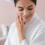 a woman at a spa in a white robe looking down and smiling touching her skin